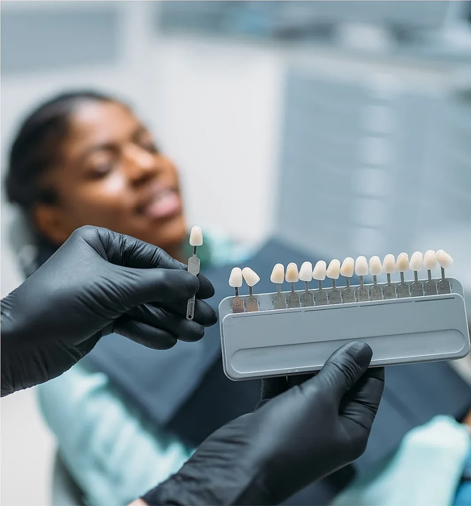 A gloved dentist's hand holds a dental shade guide against a blurred background of a smiling patient in a dental chair