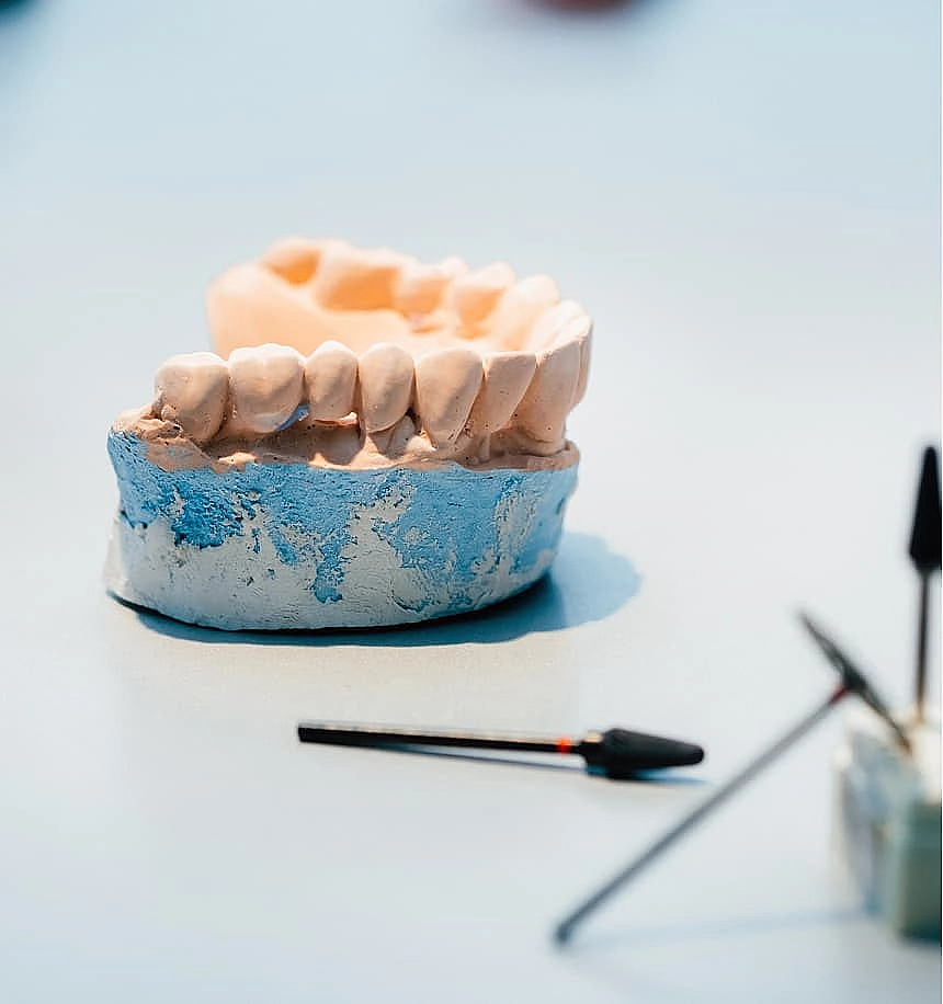 Dental model of an upper jaw with realistic teeth on a blue base. Nearby, dental tools rest on a pale surface, suggesting a clinical setting.