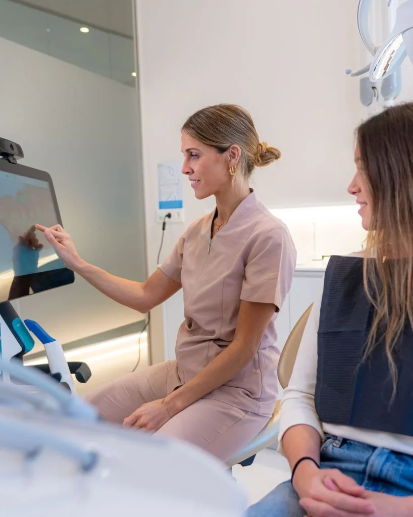 A female dental professional in light pink scrubs sits next to a patient and points to a digital 3D dental scan of teeth on a touchscreen monitor.