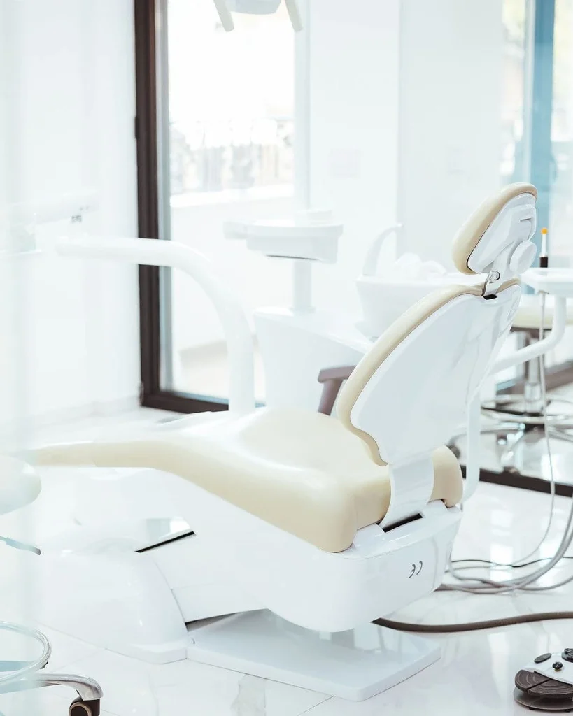 A modern, clean dental treatment room featuring an empty, high-tech white dental chair with cream-colored leather cushioning, surrounded by professional medical equipment.