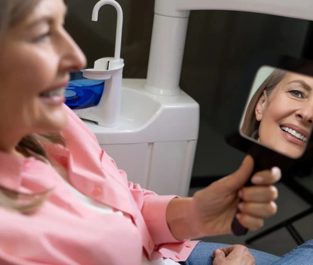 Smiling woman in a dental clinic looking at her healthy smile in a handheld mirror after cosmetic or restorative dental treatment.