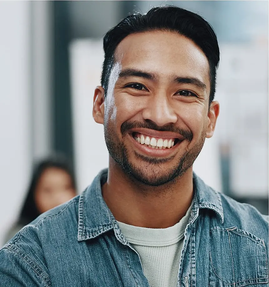 Smiling man in a denim shirt, close-up. Blurred background suggests an indoor setting, conveying warmth and positivity.