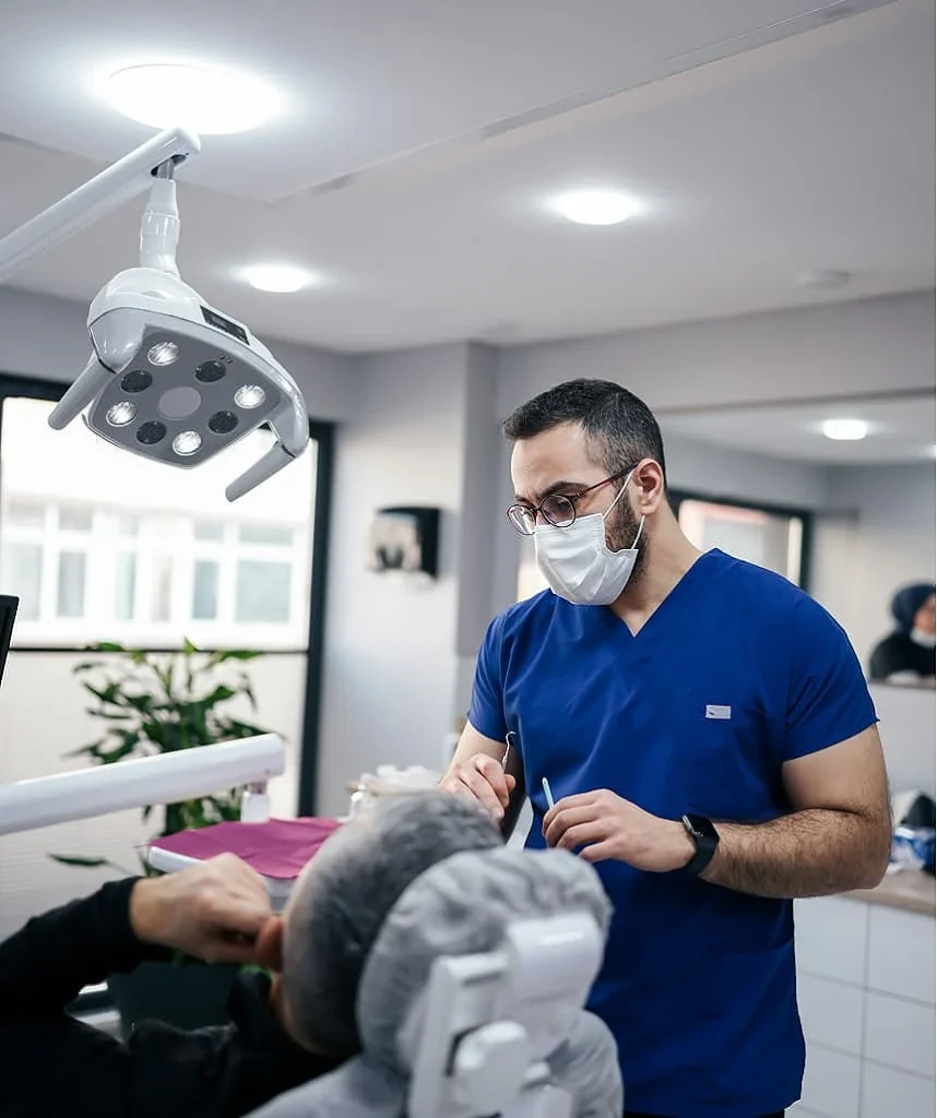 A dentist in blue scrubs and a mask examines a patient in a dental office. Bright overhead light illuminates the scene, creating a professional atmosphere.