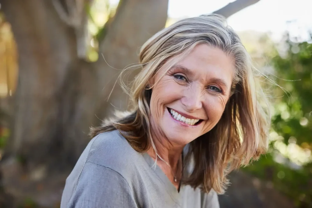 Smiling mature woman with healthy skin and shoulder-length blonde hair outdoors beneath a large tree, showcasing natural beauty and healthy aging.