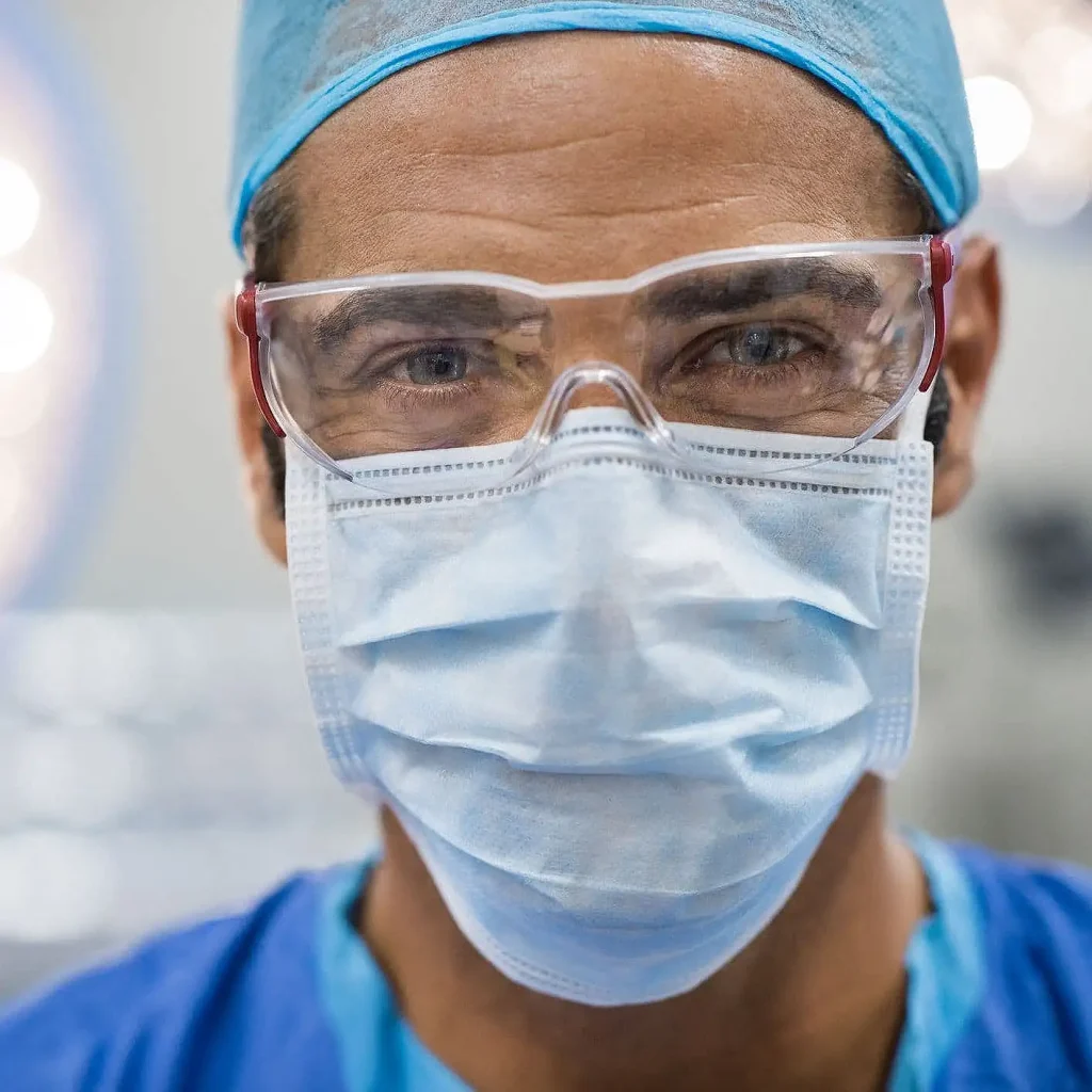 Smiling healthcare professional wearing a blue surgical mask, goggles, and cap, exuding calm and focus in a clinical setting.