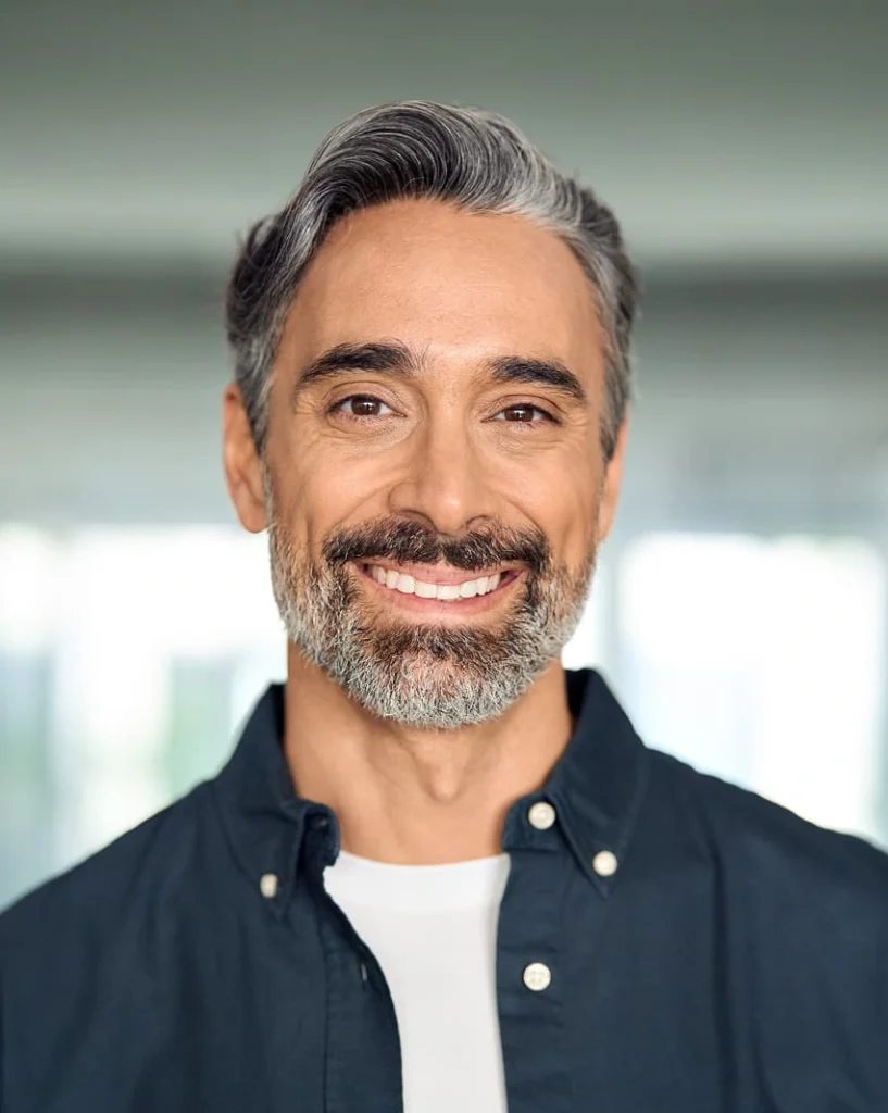 Smiling man with gray hair and beard, wearing a dark shirt over a white t-shirt, standing indoors with a blurred background, conveying a friendly and warm atmosphere.
