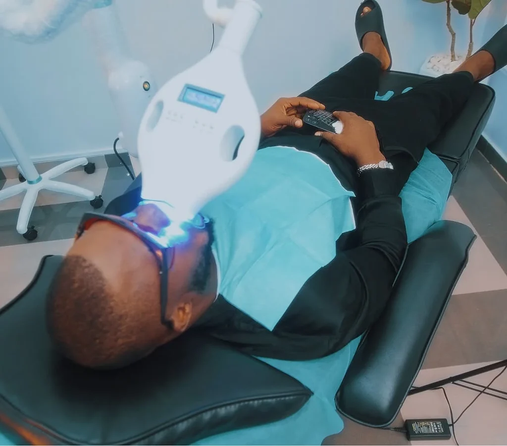 A person lies on a reclining dental chair undergoing teeth whitening, wearing protective eyewear. A bright blue light shines on their mouth.