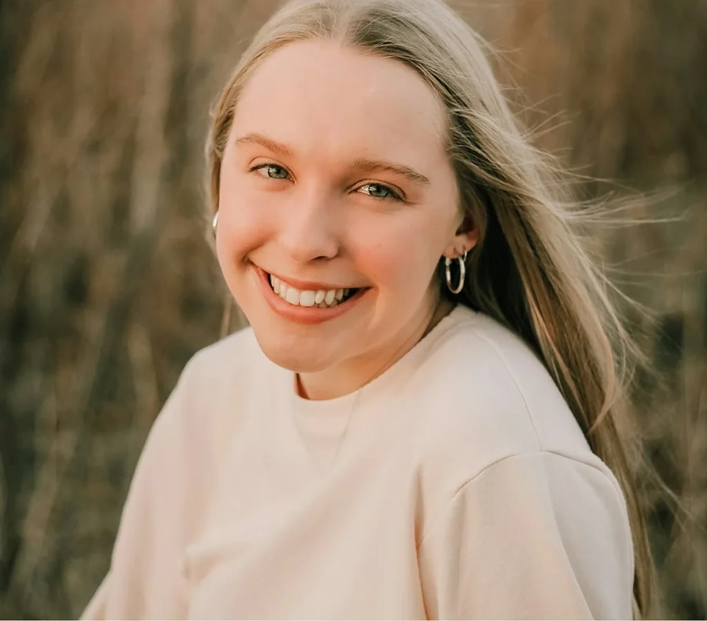 Smiling woman with long blonde hair and hoop earrings, wearing a light top. Her expression is joyful against a blurred, natural outdoor background.