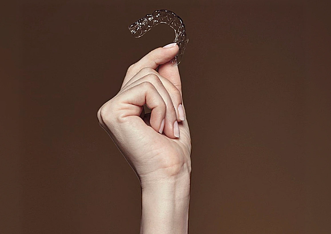 Hand holding a clear removable dental aligner against a solid brown background