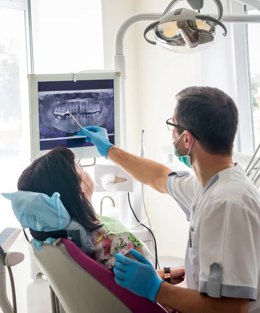 A dentist pointing with a tool at a panoramic dental X-ray on a monitor while explaining the diagnosis to a patient.