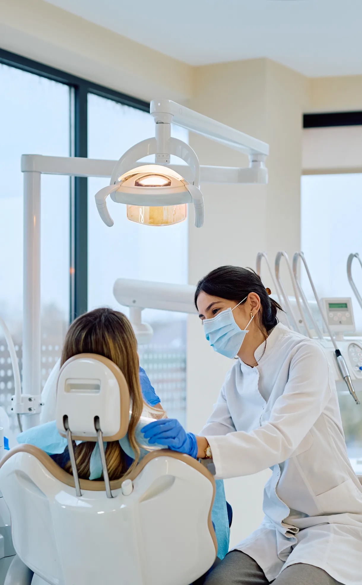 A female dentist wearing a white coat, surgical mask, and blue gloves attending to a patient sitting in a dental chair.