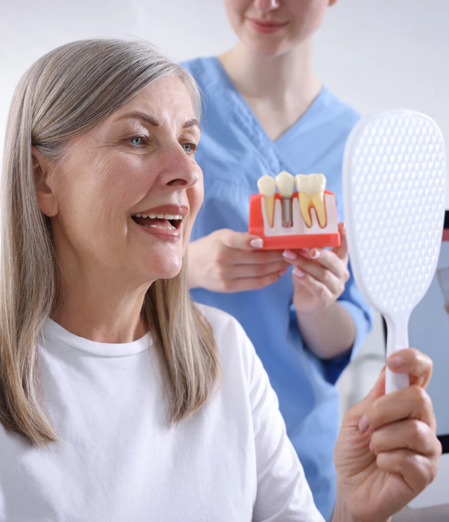 Smiling senior woman looking in a mirror after a dental implant consultation while a dentist demonstrates an implant model, highlighting restored smiles and improved oral health.